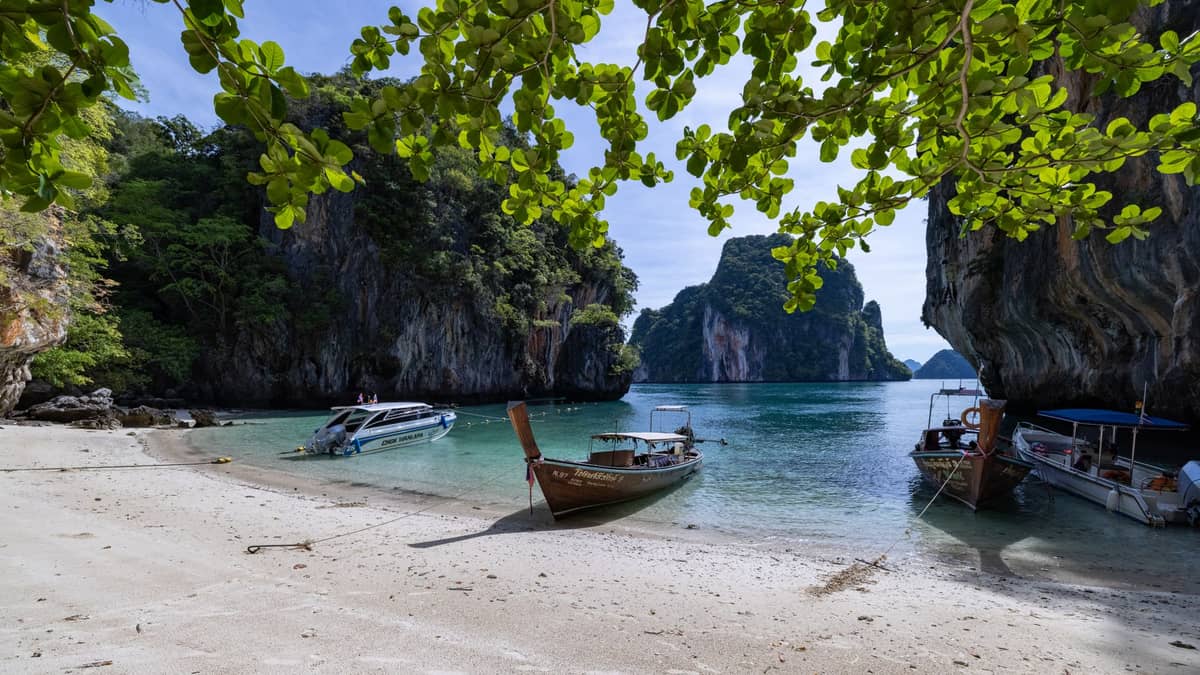 Botes de cola larga y una lancha rápida anclados en una playa de arena en Krabi, rodeados de imponentes acantilados de piedra caliza y agua turquesa bajo árboles frondosos.