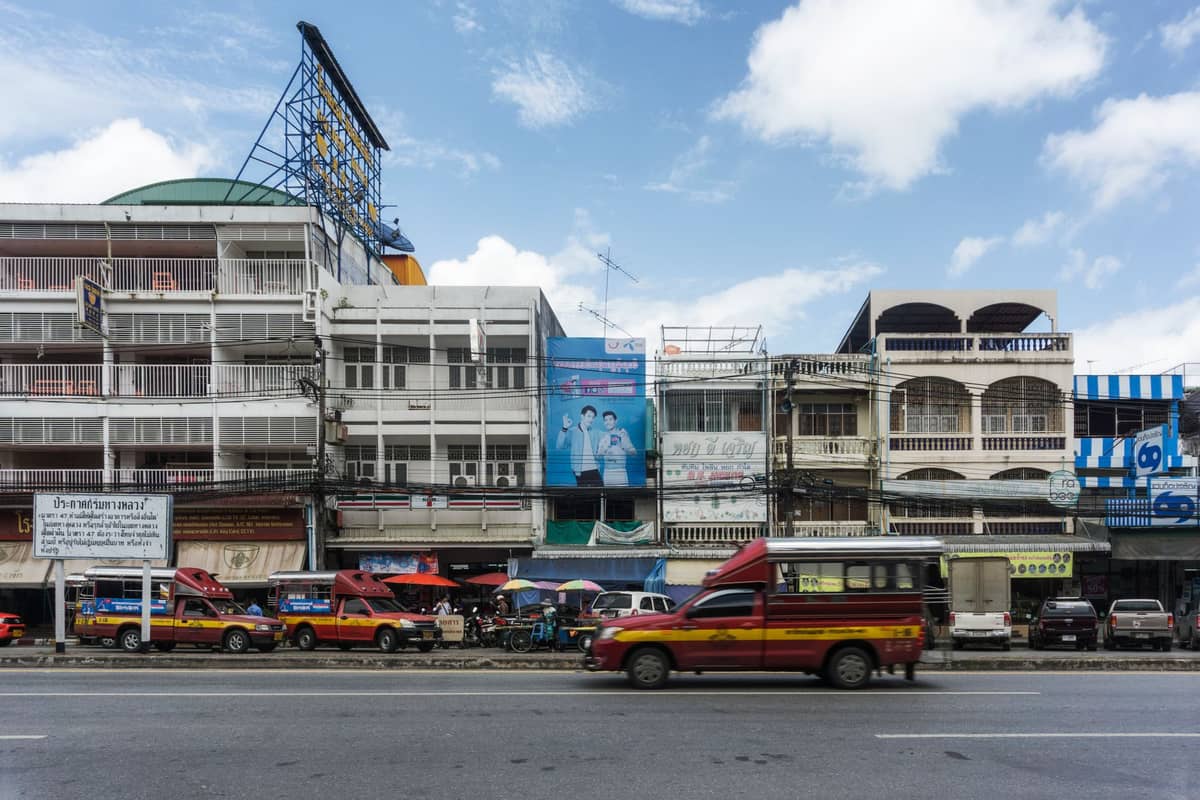 Fila de songthaews rojos estacionados en una calle frente a tiendas y apartamentos tailandeses bajo un cielo azul
