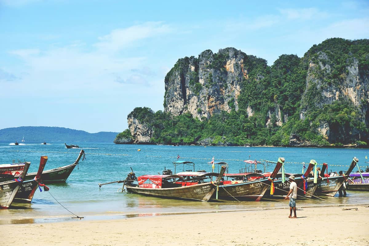Varios botes de cola larga tradicionales alineados a lo largo de una playa de arena con dramáticos acantilados de piedra caliza y mar azul en Krabi, Tailandia.