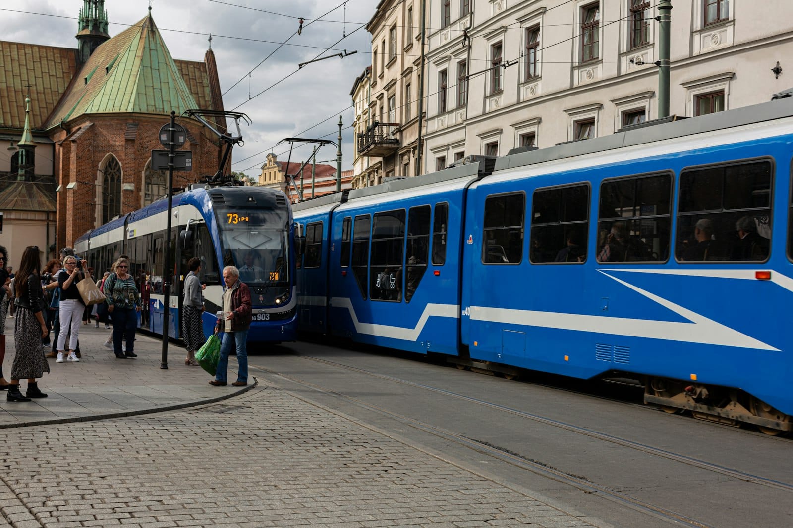 รถรางสีน้ำเงิน MPK Kraków พร้อมผู้โดยสารกำลังขึ้น-ลงที่ป้ายบนถนนในเมือง รายล้อมด้วยอาคารประวัติศาสตร์