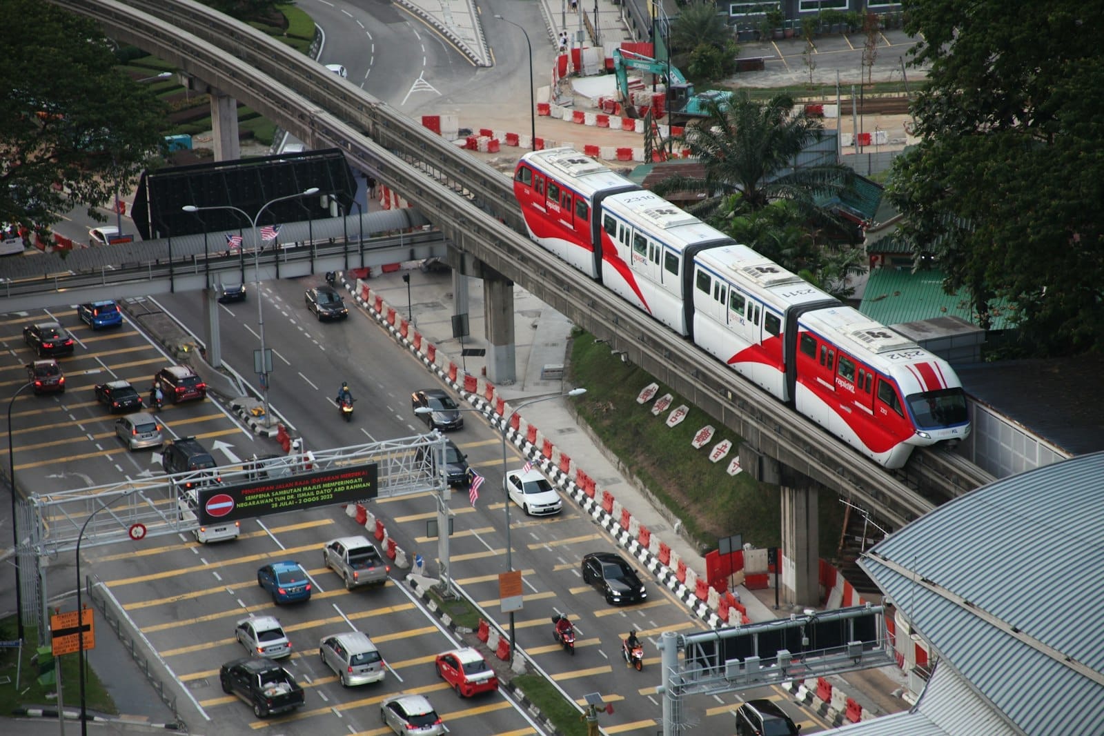 Aerial view of the Kuala Lumpur Monorail traveling above busy city streets filled with cars and motorcycles, surrounded by urban greenery and buildings.