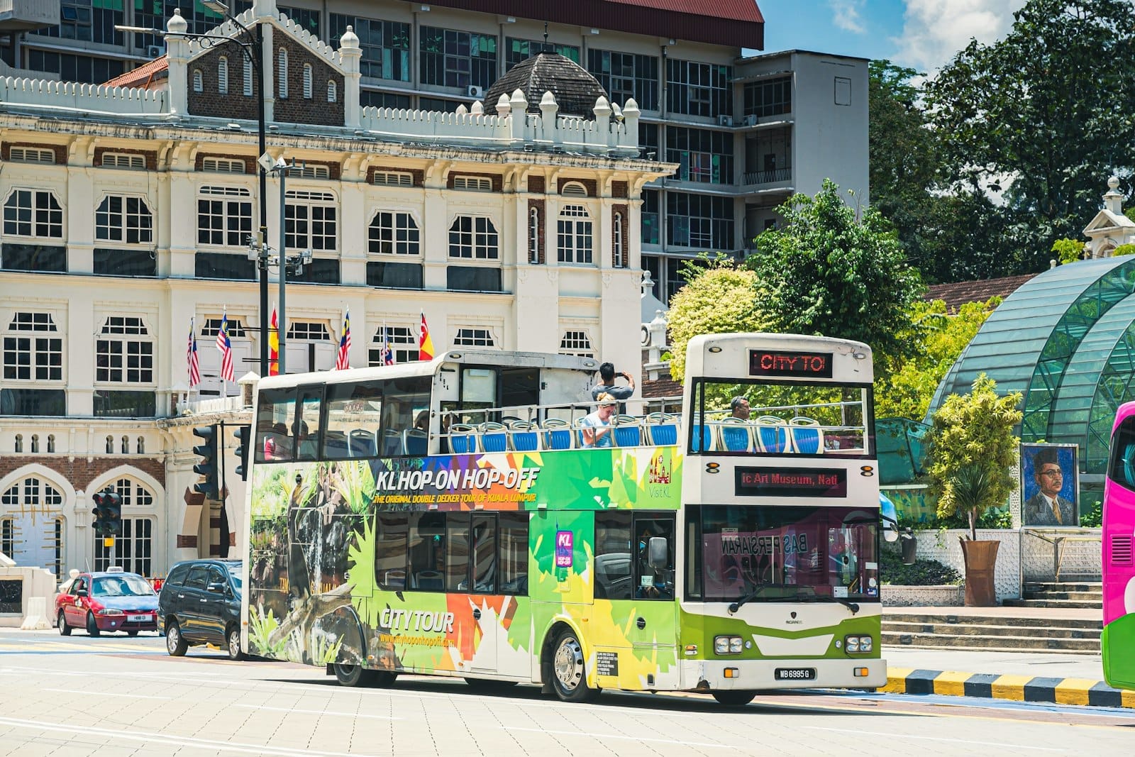 Colorful Kuala Lumpur hop-on hop-off double-decker bus passing colonial buildings and Malaysian flags on a sunny city street.