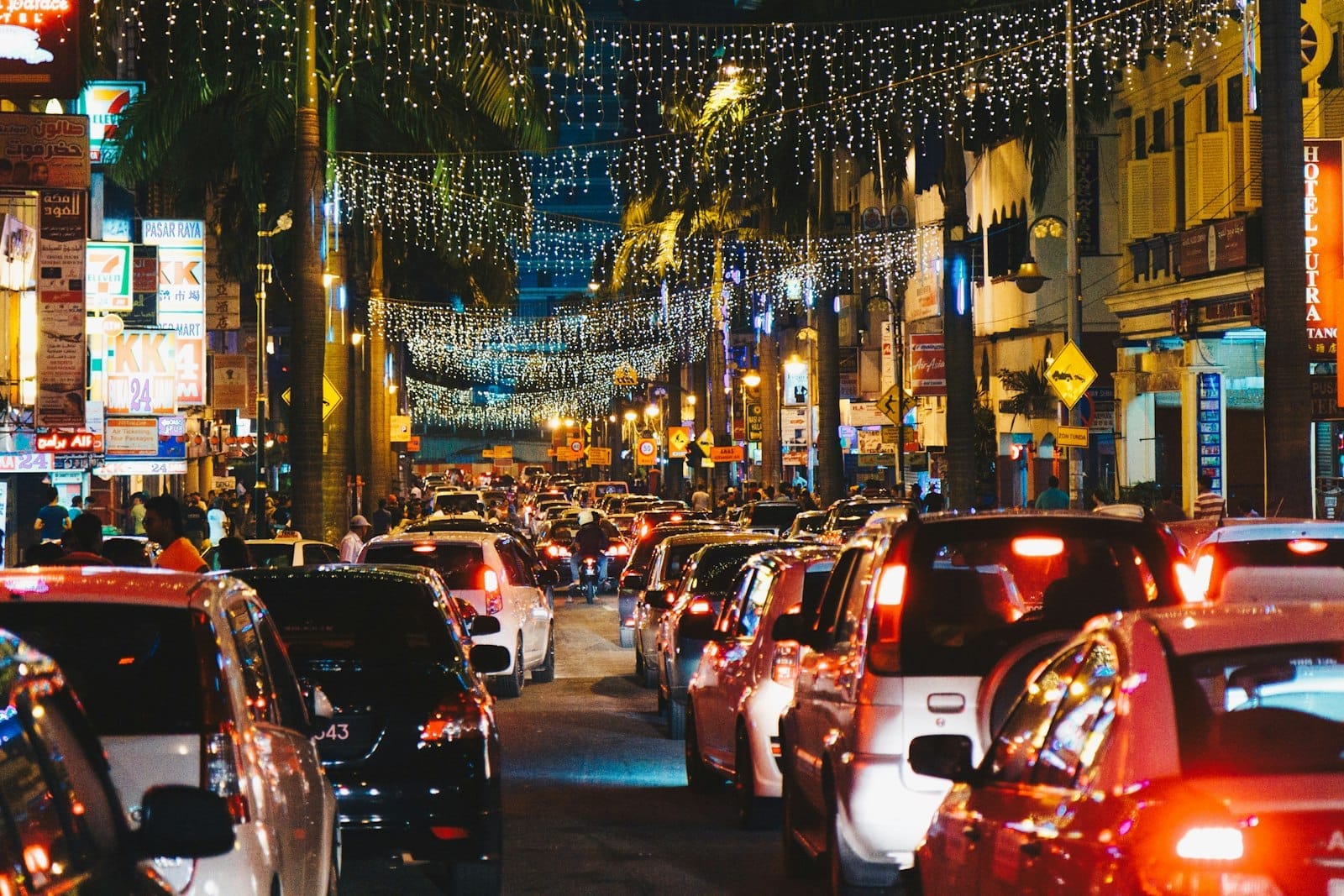 Heavy evening traffic fills a brightly lit street in Kuala Lumpur, with colorful shop signs and festive string lights spanning above busy cars.