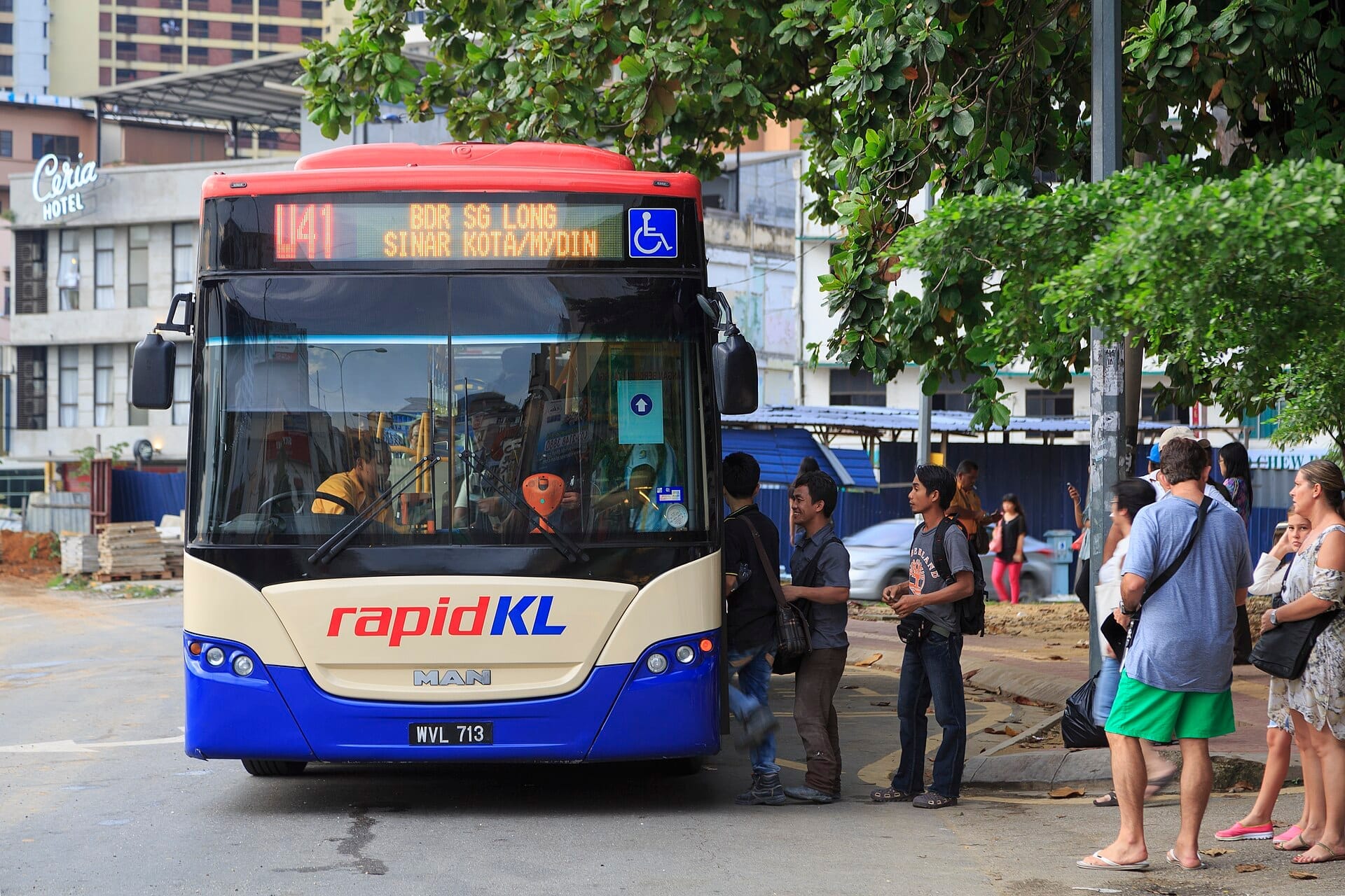 People queuing to board a RapidKL city bus under leafy trees in central Kuala Lumpur, with buildings and urban construction in the background.