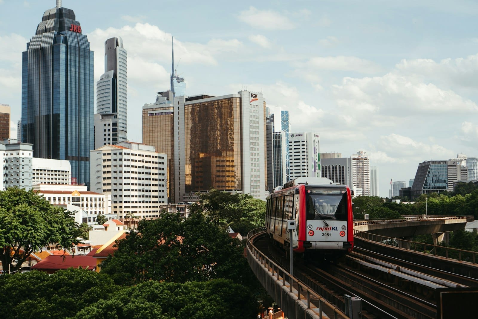 Rapid KL train traveling on elevated tracks with Kuala Lumpur city skyline, modern skyscrapers, and lush green trees under a partly cloudy sky.