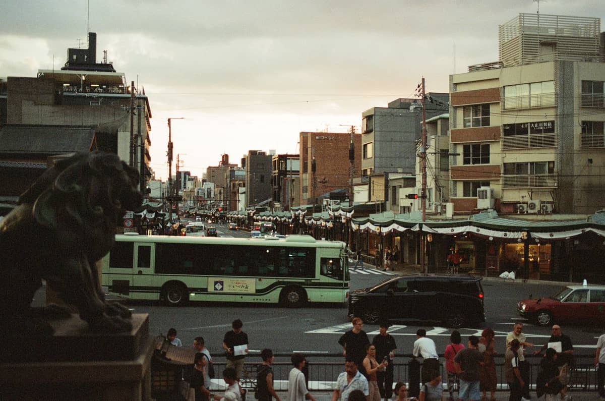 A Kyoto city bus driving through a busy street lined with traditional low-rise buildings and many pedestrians at dusk.