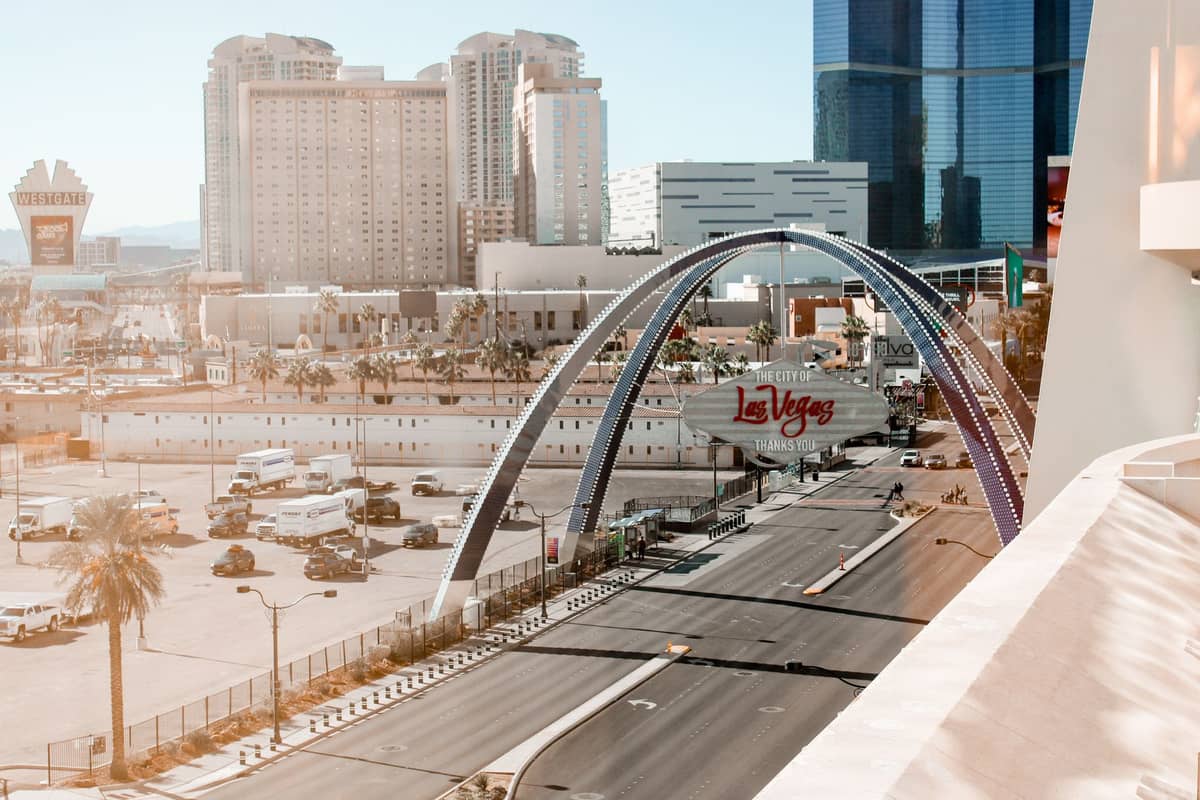 Una calle de la ciudad en Las Vegas con coches y un prominente cartel de 'Las Vegas' arqueado sobre la carretera, rodeado de hoteles modernos y palmeras.