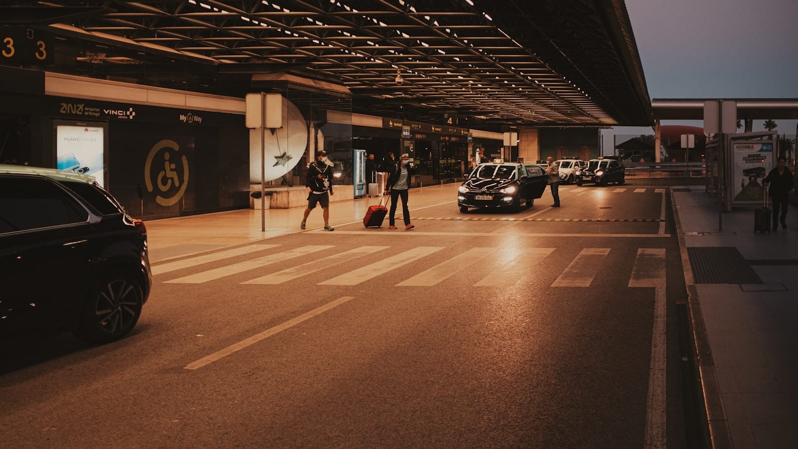 Carros e viajantes na entrada de um terminal moderno do aeroporto de Lisboa ao entardecer.