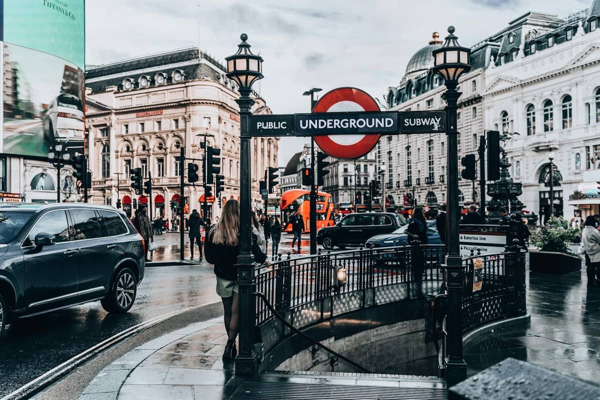 Entrada de uma estação do metrô de Londres ao nível da rua em Piccadilly Circus, com o icônico sinal roundel e uma movimentada cena urbana durante o dia.