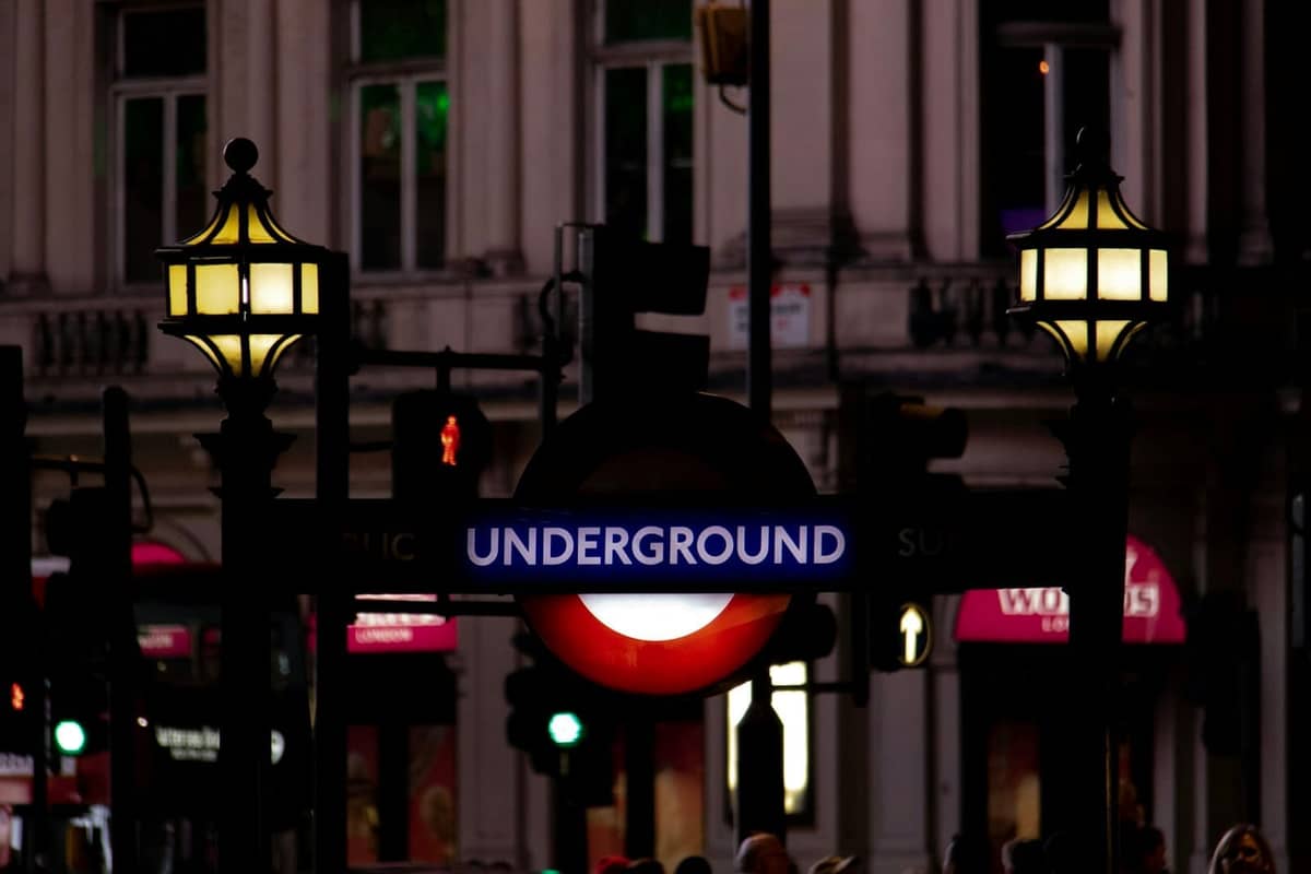Icônico letreiro de entrada do London Underground com lâmpadas brilhantes à noite, em frente à fachada de um histórico edifício da cidade.