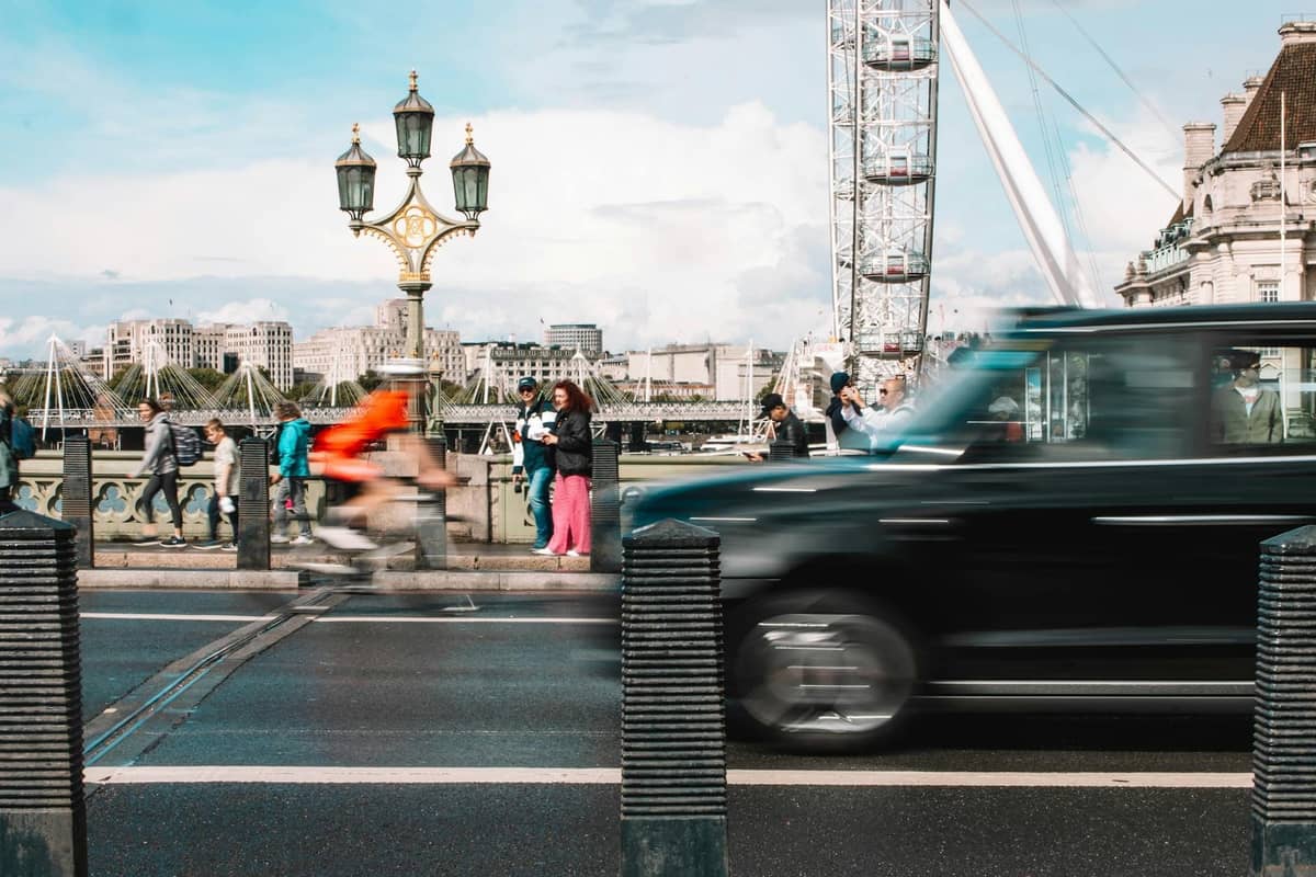 Cena de rua em Londres com um táxi preto, um ciclista em movimento e o London Eye ao fundo numa ponte movimentada.