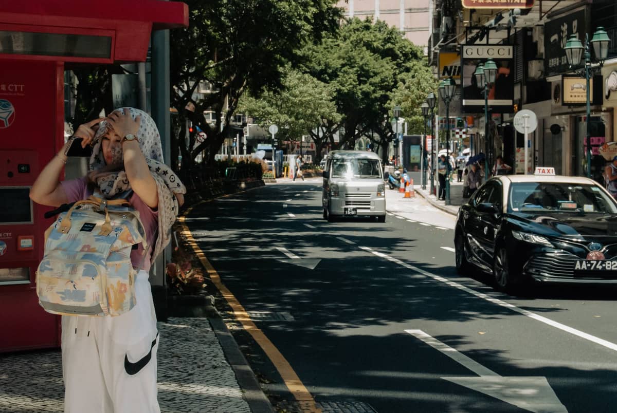 Ein schwarzes Taxi fährt durch eine Straße im Zentrum von Macau mit lokalen Geschäften und einem Fußgänger in der Nähe.