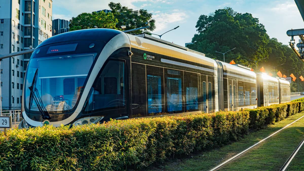 Moderner LRT-Zug in Macau an einem Außenbahnhof, umgeben von Grünflächen und mittelhohen Gebäuden, mit Sonnenlichtreflexen auf seiner eleganten Außenhülle.