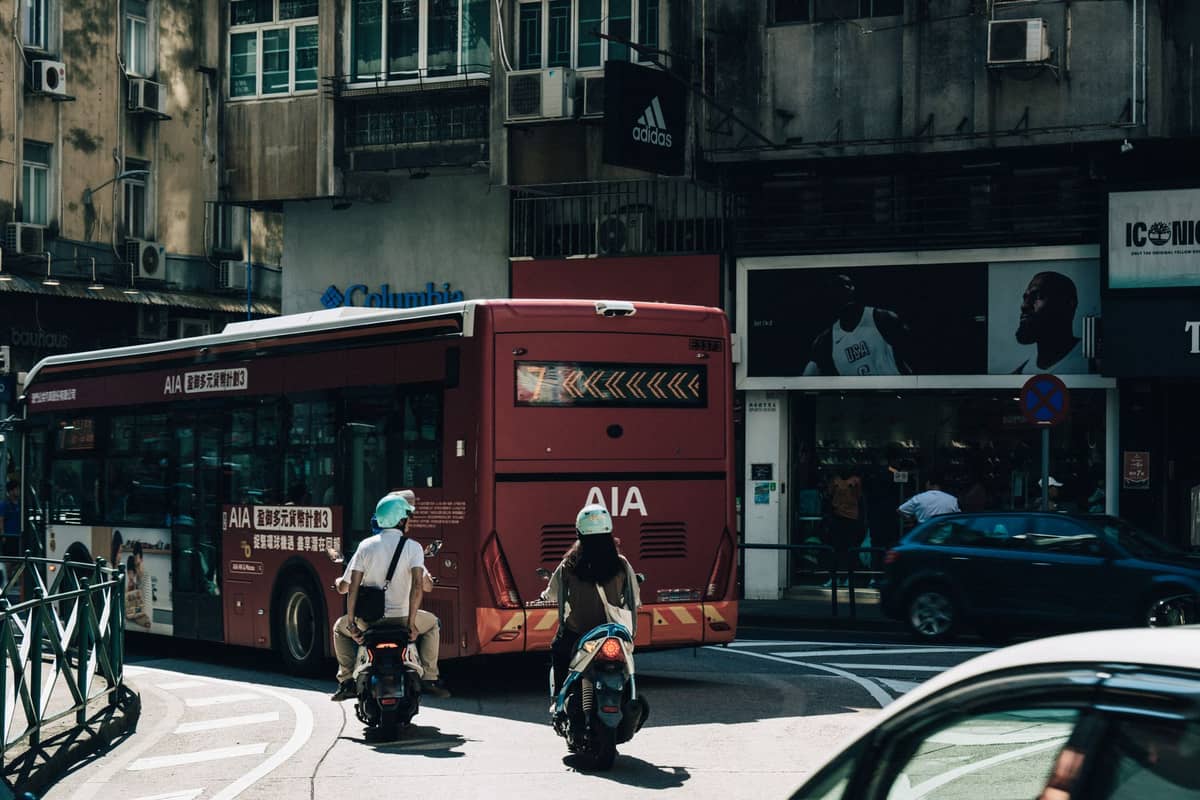 Roter öffentlicher Bus mit zweisprachiger Beschilderung auf einer belebten Stadtstraße in Macau, Motorräder in der Nähe und Geschäfte im Hintergrund.