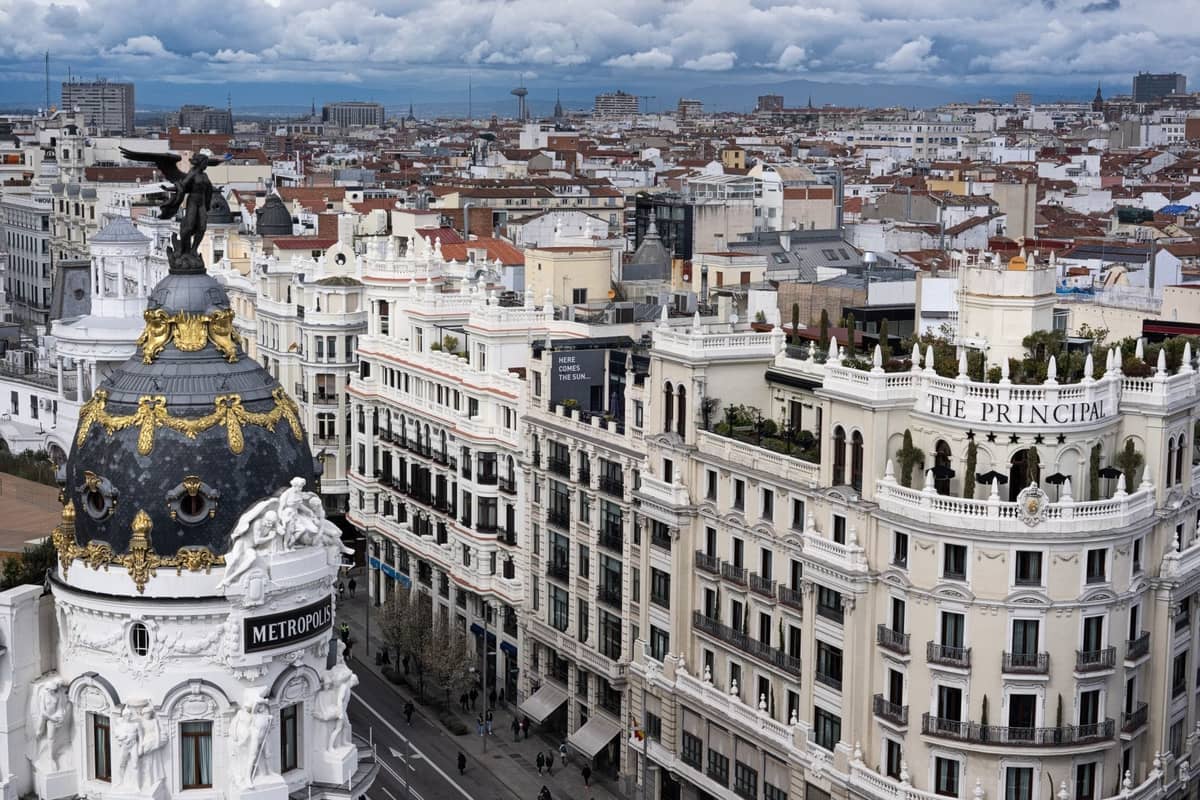 Luftaufnahme der Gran Vía in Madrid mit dem ikonischen Metropolis-Gebäude, belebten Straßen und Stadtpanorama unter bewölktem Himmel.