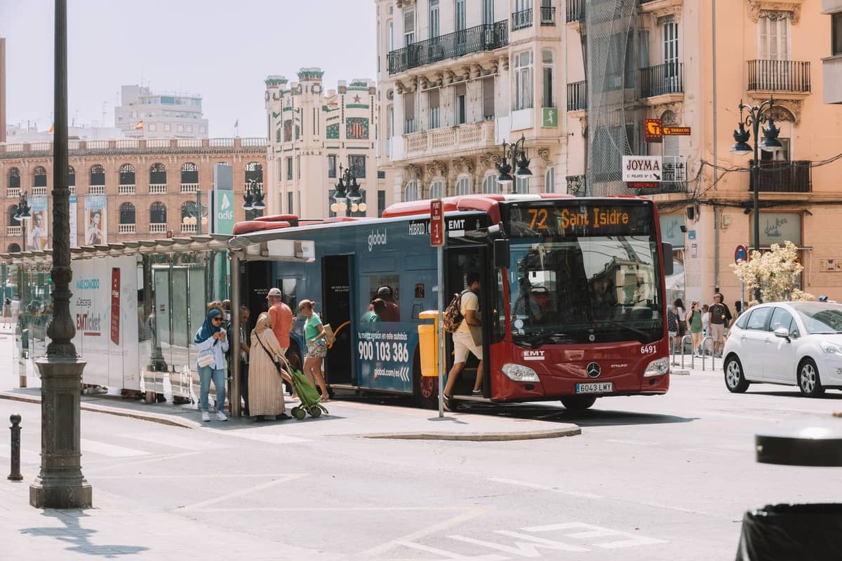 Roter EMT Madrid-Stadtbus mit einsteigenden Fahrgästen an einer Haltestelle in einer belebten Straße in Madrid, mit klassischen Gebäuden im Hintergrund.