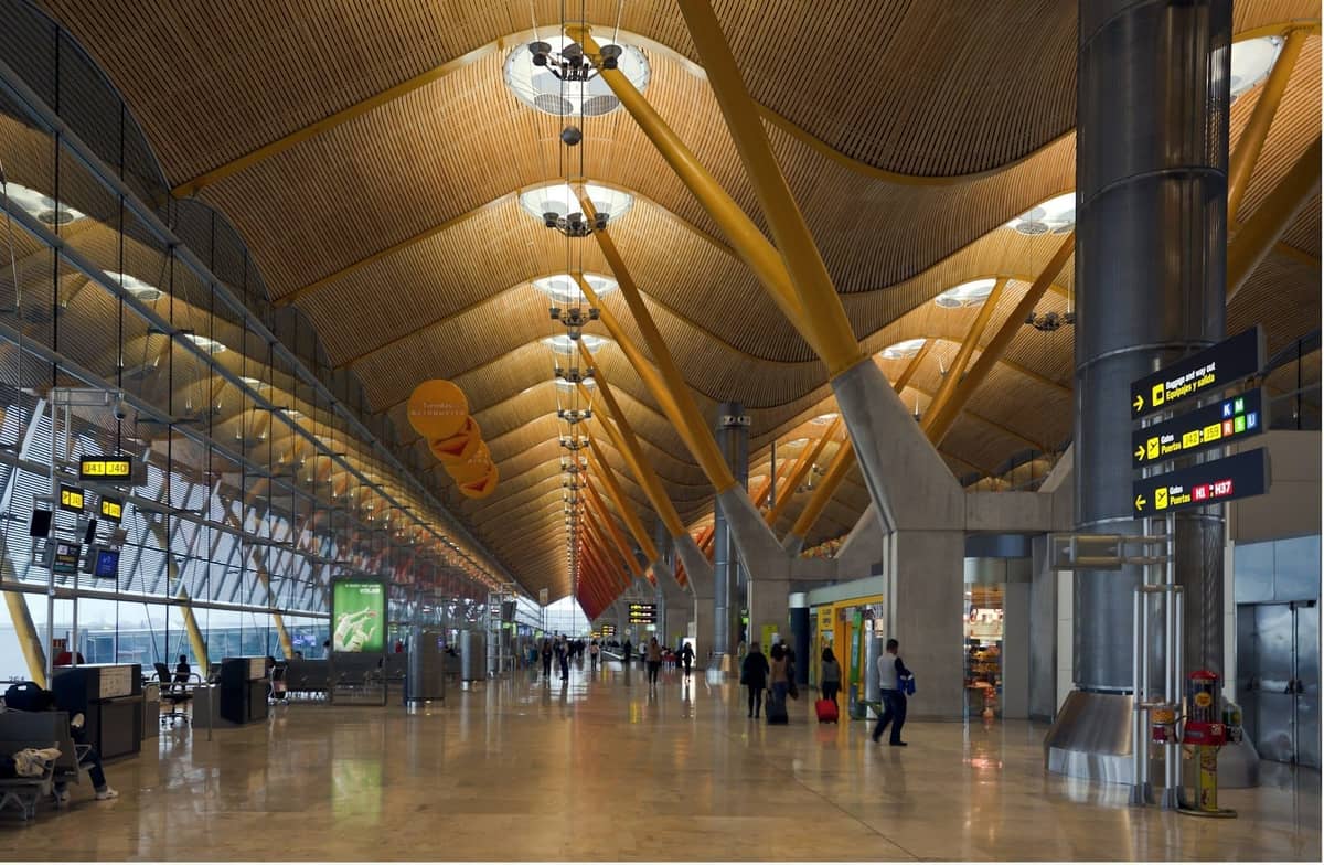 Geräumiges Inneres des Terminals des Flughafens Madrid Barajas mit gelben Trägern, Beschilderung und Reisenden, die durch die Halle laufen.