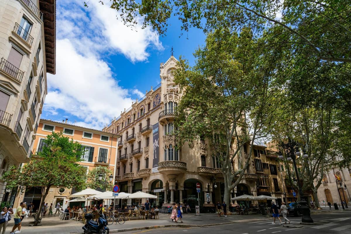 Trung tâm Palma de Mallorca với các quán cà phê ngoài trời, người qua lại, các tòa nhà lịch sử và cây xanh tươi tốt tại một ngã tư đô thị sầm uất.