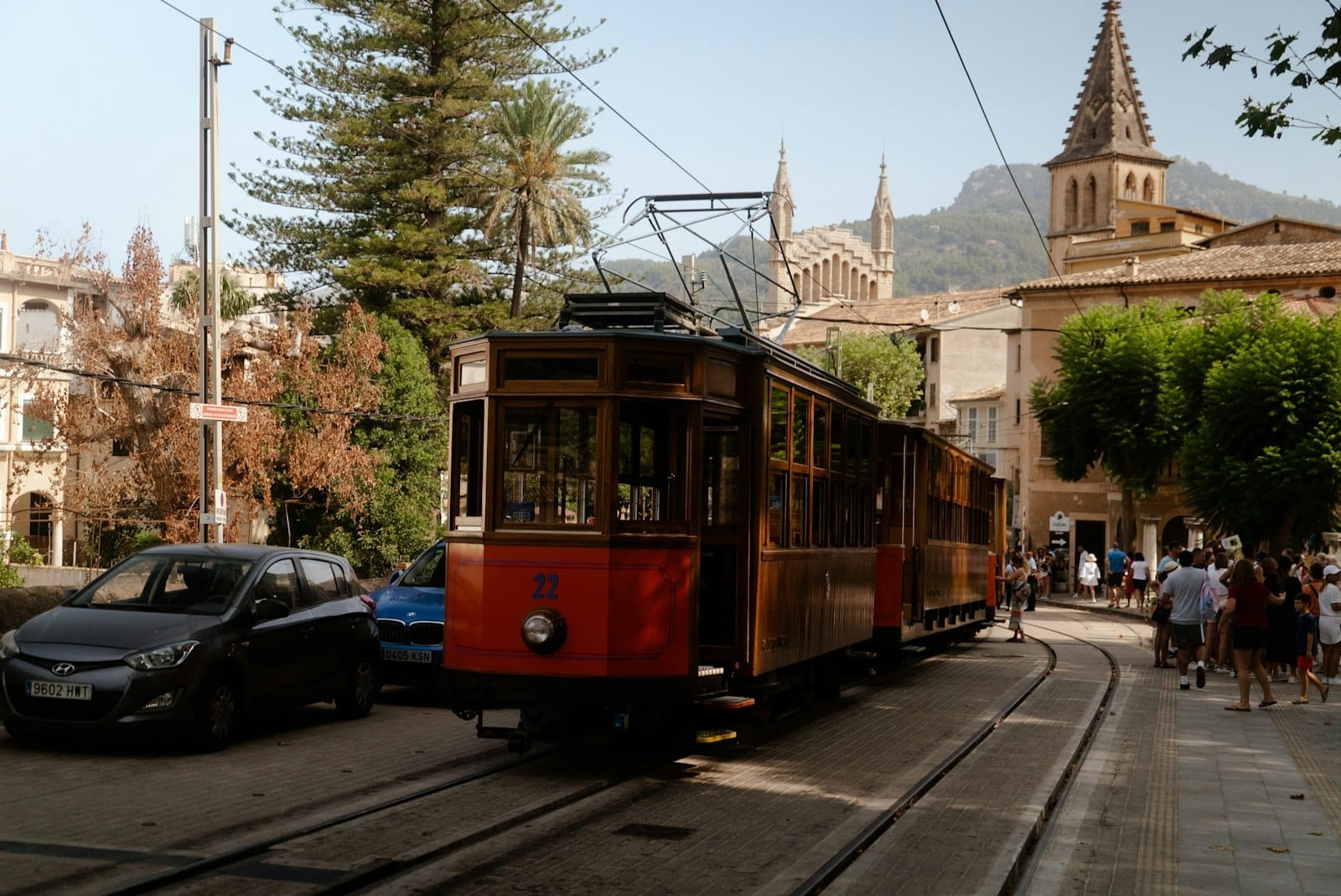 Bonde de madeira vintage passando por uma cidade pitoresca com carros e pessoas em Sóller, Maiorca, com montanhas e uma igreja ao fundo.