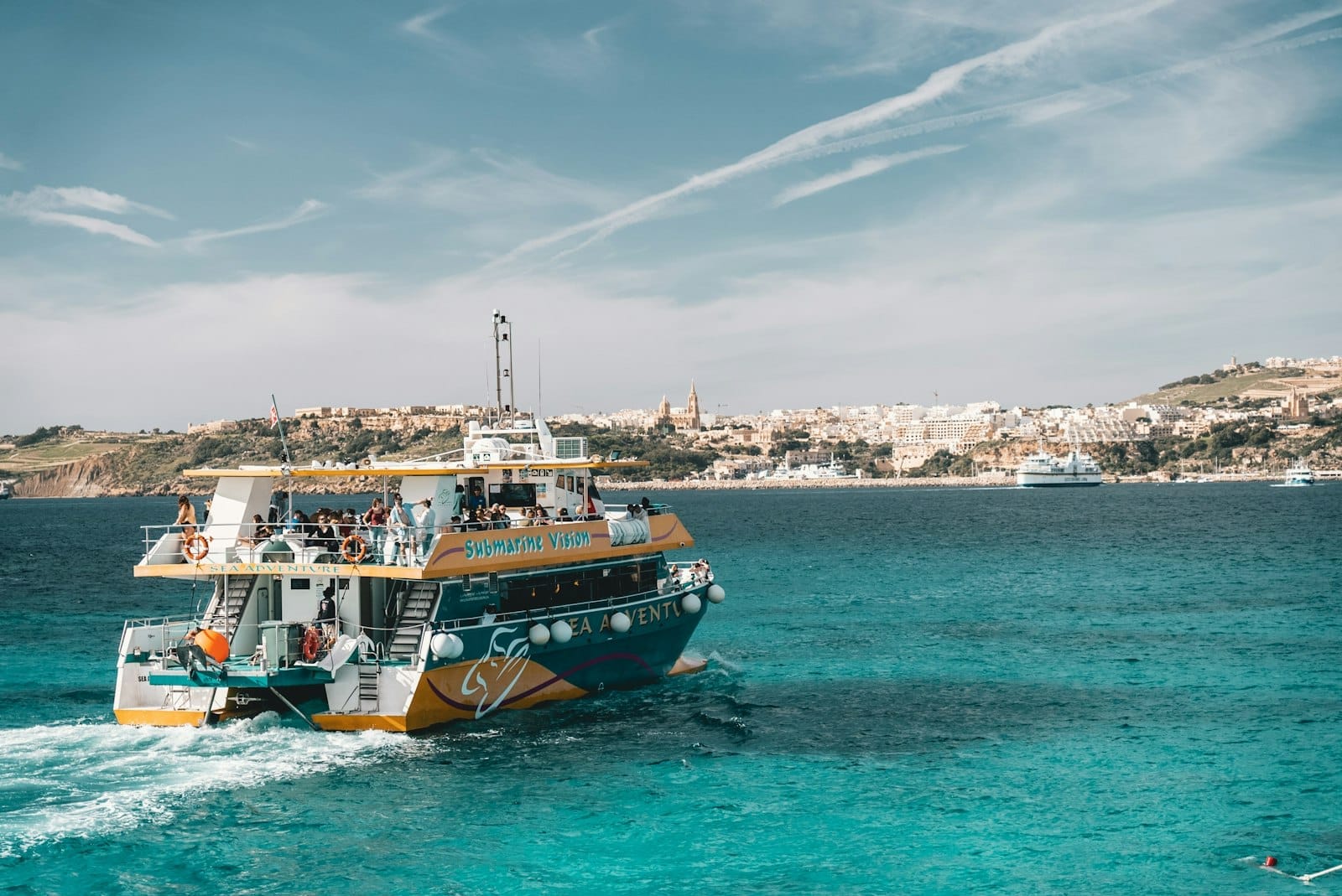 Uma balsa colorida com passageiros cruza águas turquesa com a costa e uma cidade maltesa ao fundo, sob um céu azul.