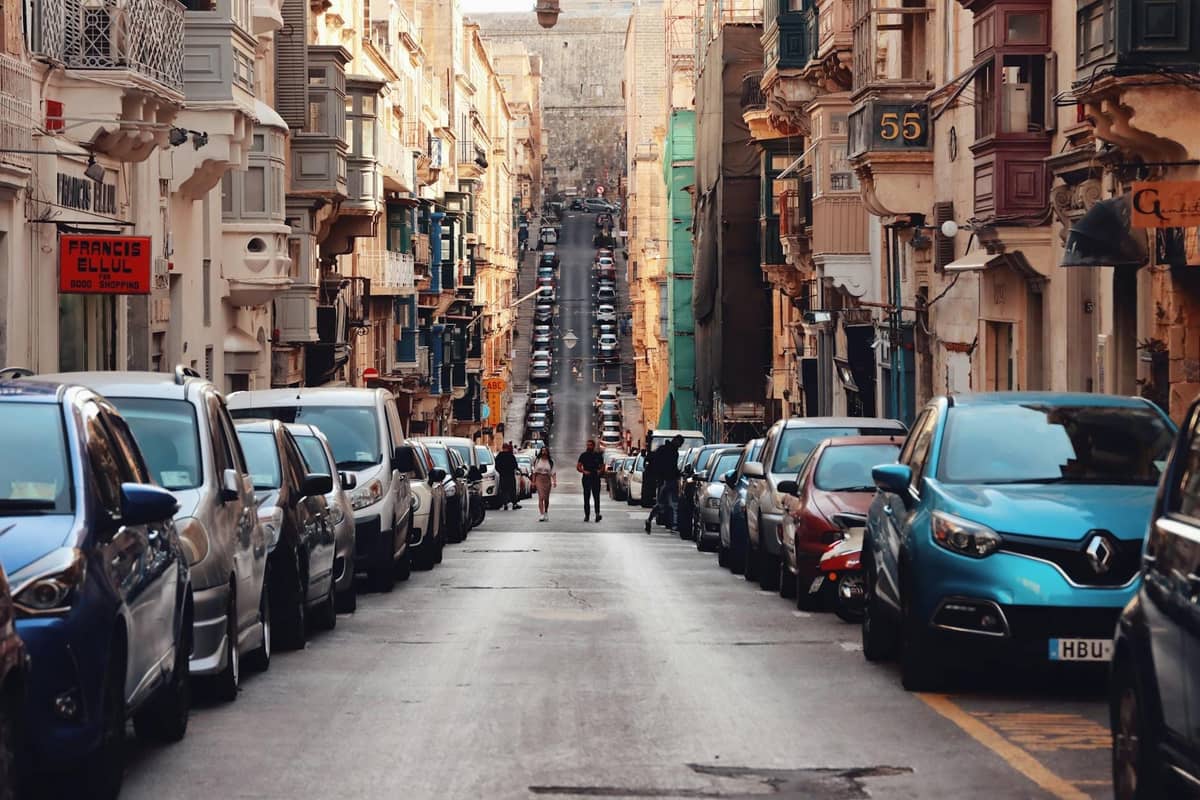 Una vivace strada maltese fiancheggiata da auto parcheggiate, edifici tradizionali e alcune persone che camminano al centro della carreggiata.
