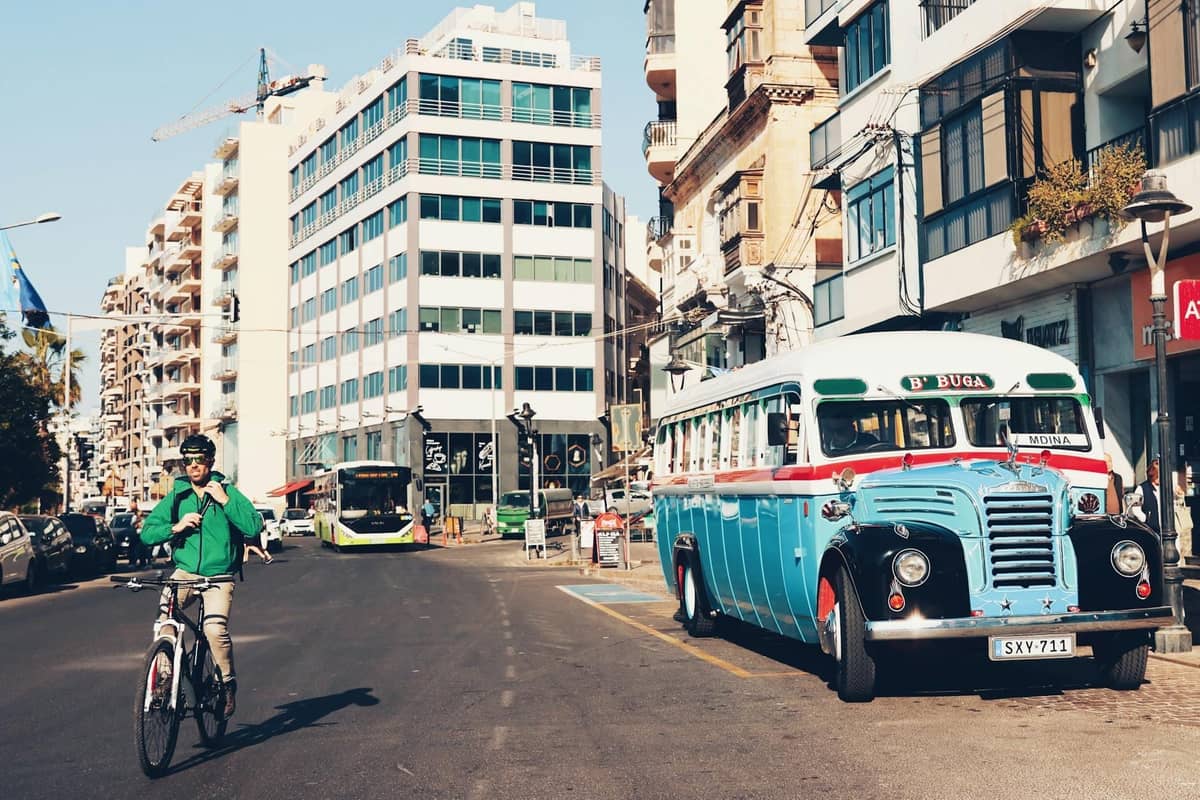 Autobus maltese classico parcheggiato in una strada della città, con edifici moderni e un ciclista in primo piano, che mostra il contesto urbano di Malta.