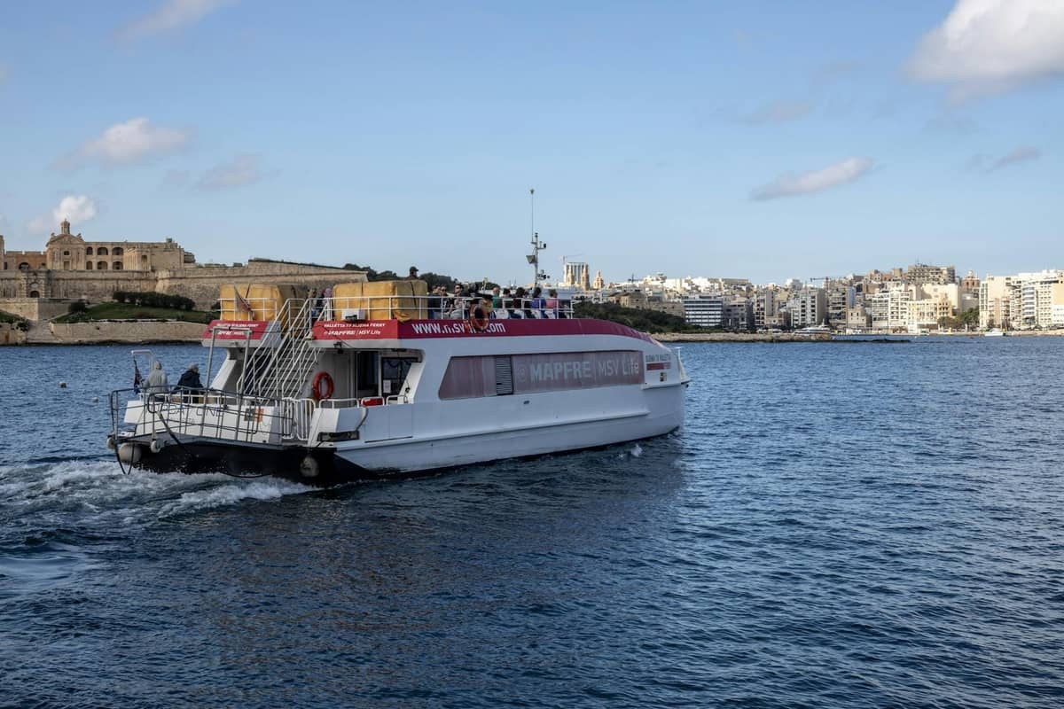 Un traghetto passeggeri che attraversa un porto a Malta con lo skyline e le fortificazioni di Valletta visibili sullo sfondo sotto un cielo azzurro.