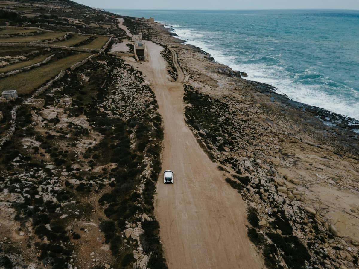 Una piccola auto percorre una strada costiera accidentata a Malta con terreno roccioso e il Mar Mediterraneo su un lato.