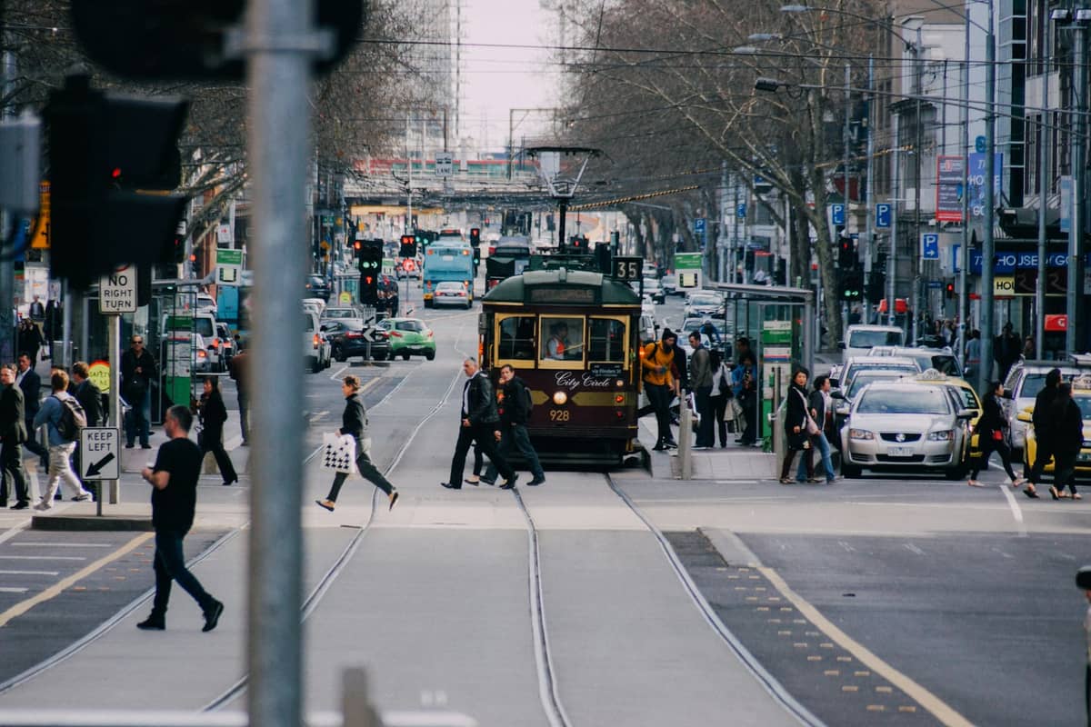 A classic Melbourne tram travels along a busy city street with people crossing and cars waiting, showcasing urban transport in Melbourne.