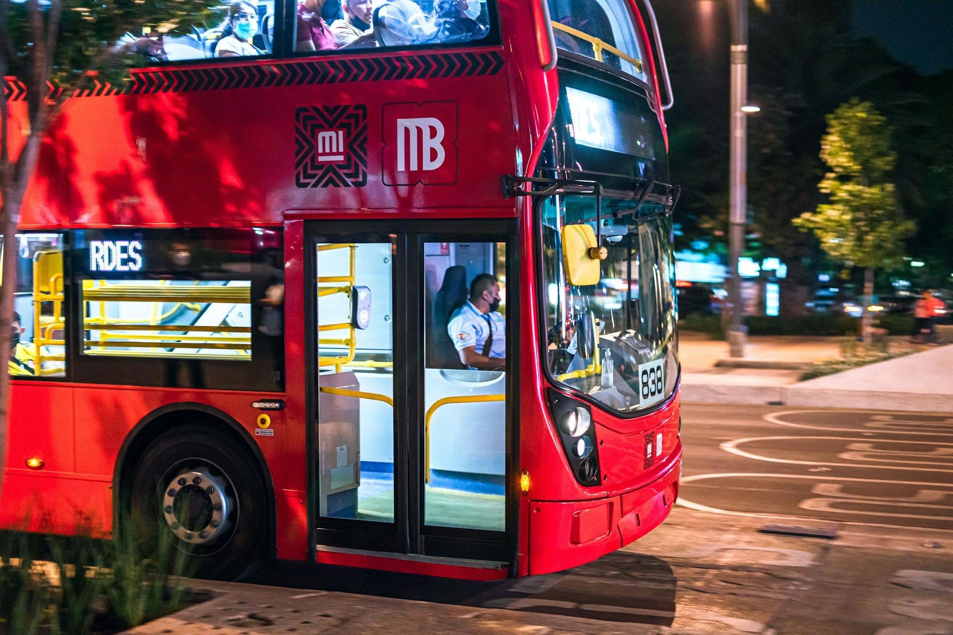 Vista de perto de um ônibus de dois andares vermelho do Metrobús à noite na Cidade do México, com passageiros visíveis pelas janelas.