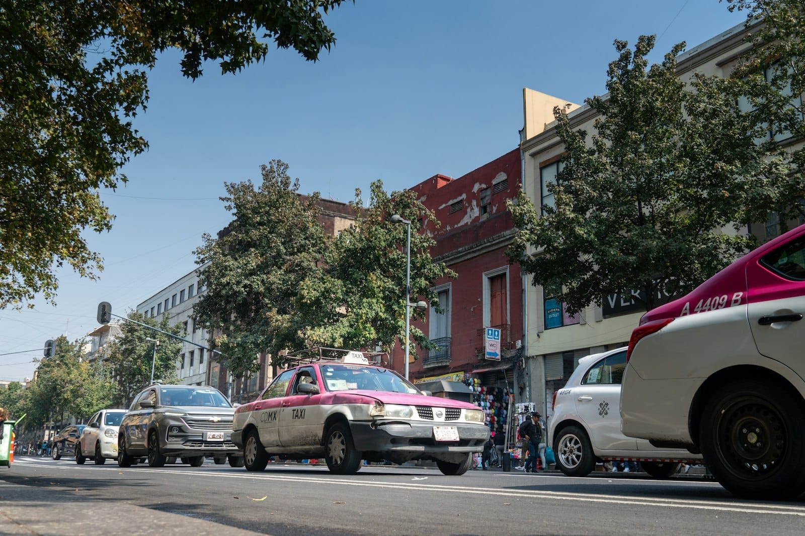 Táxis rosa e branco da CDMX e outros veículos trafegando por uma avenida central com edifícios e árvores típicos da Cidade do México ao fundo.