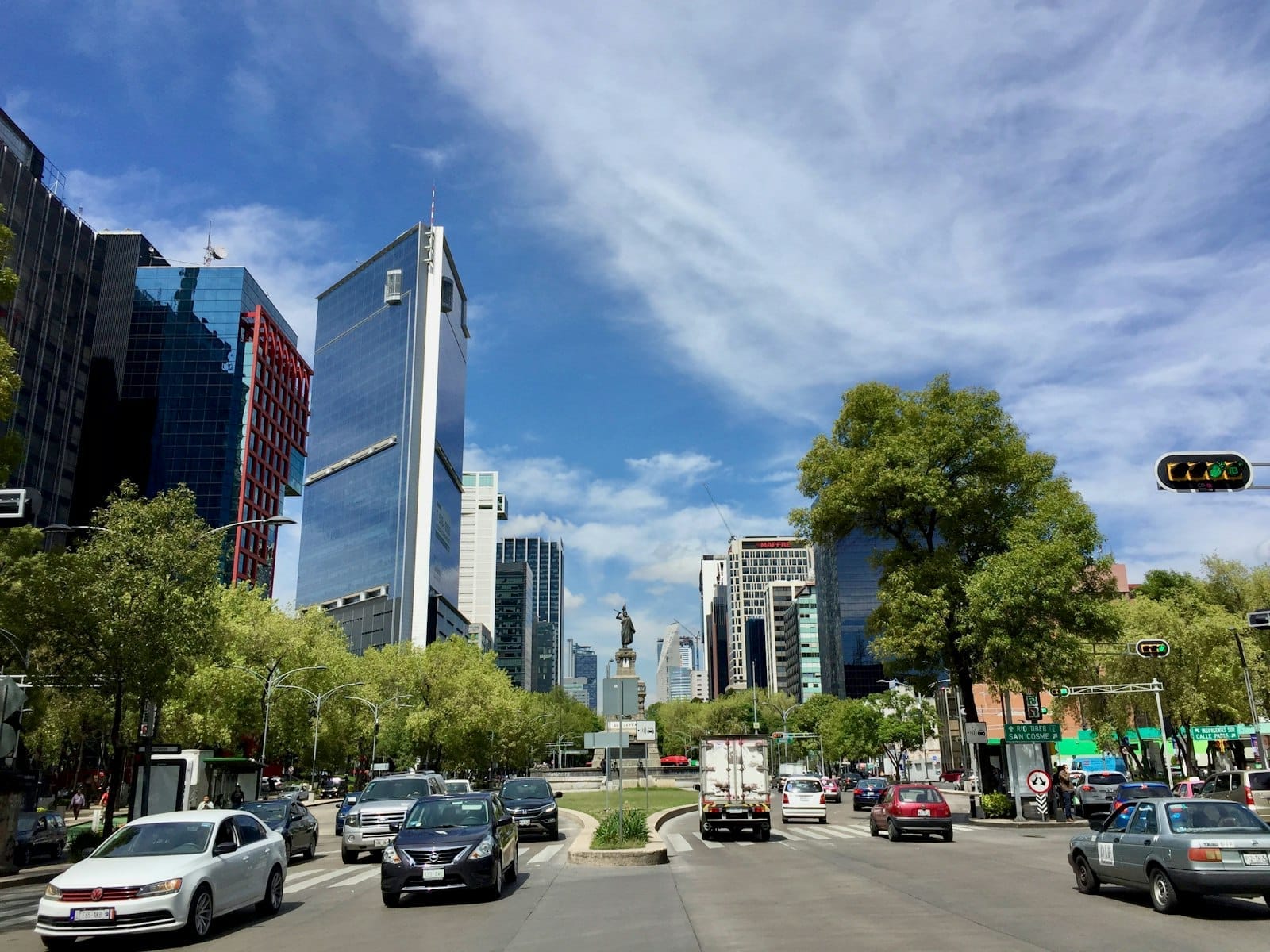 Avenida movimentada na Cidade do México com prédios modernos, carros, semáforos e o monumento do Anjo da Independência ao fundo sob um céu azul.