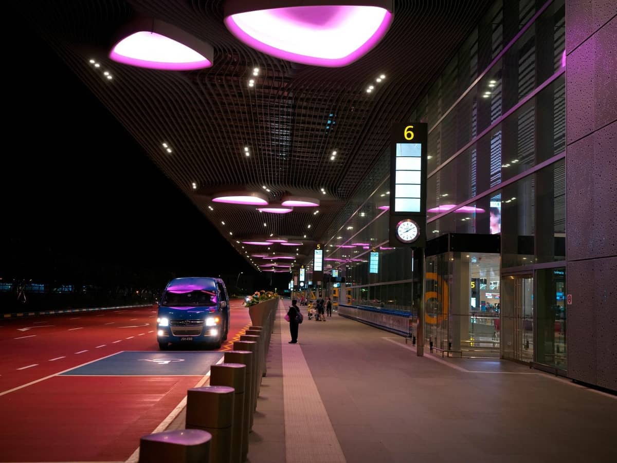 Nighttime view of an airport terminal curbside with a bus, illuminated purple ceiling lights, glass facade, and arrivals area.