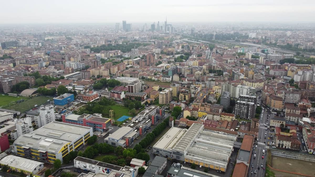 Vista panoramica aerea su Milano che mostra quartieri, strade e lo skyline in lontananza con un mix di edifici e aree verdi.