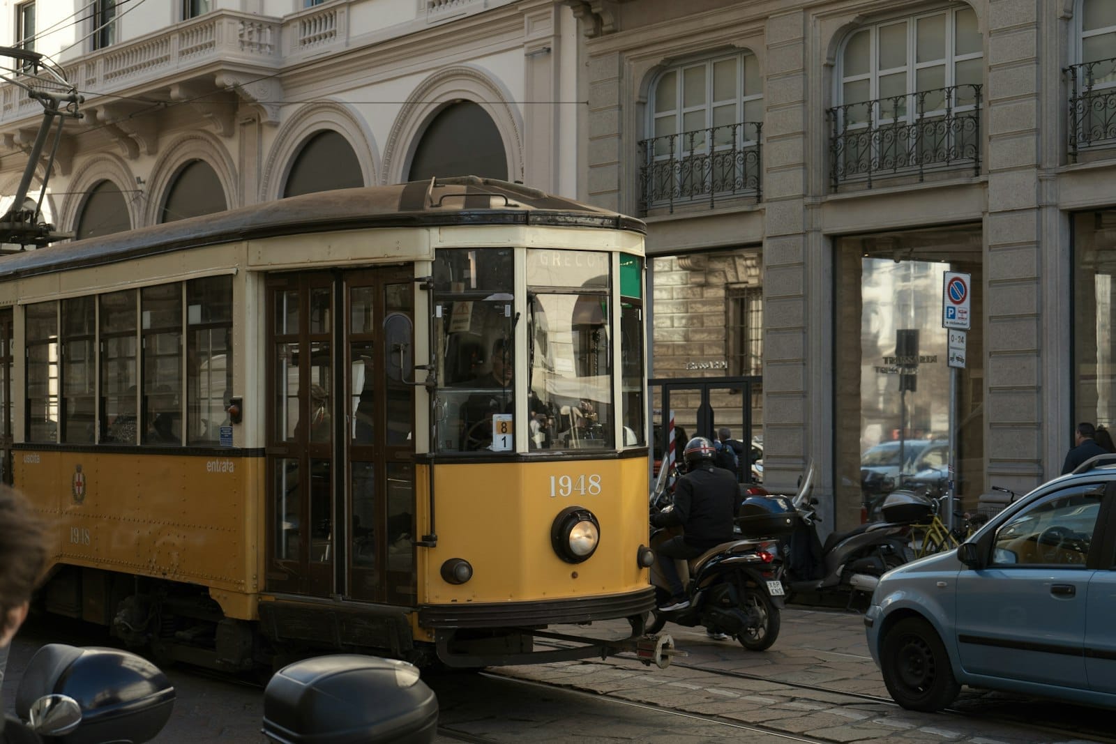 Bonde amarelo clássico de Milão percorrendo uma rua da cidade cercada por prédios históricos e scooters estacionadas em um dia ensolarado.