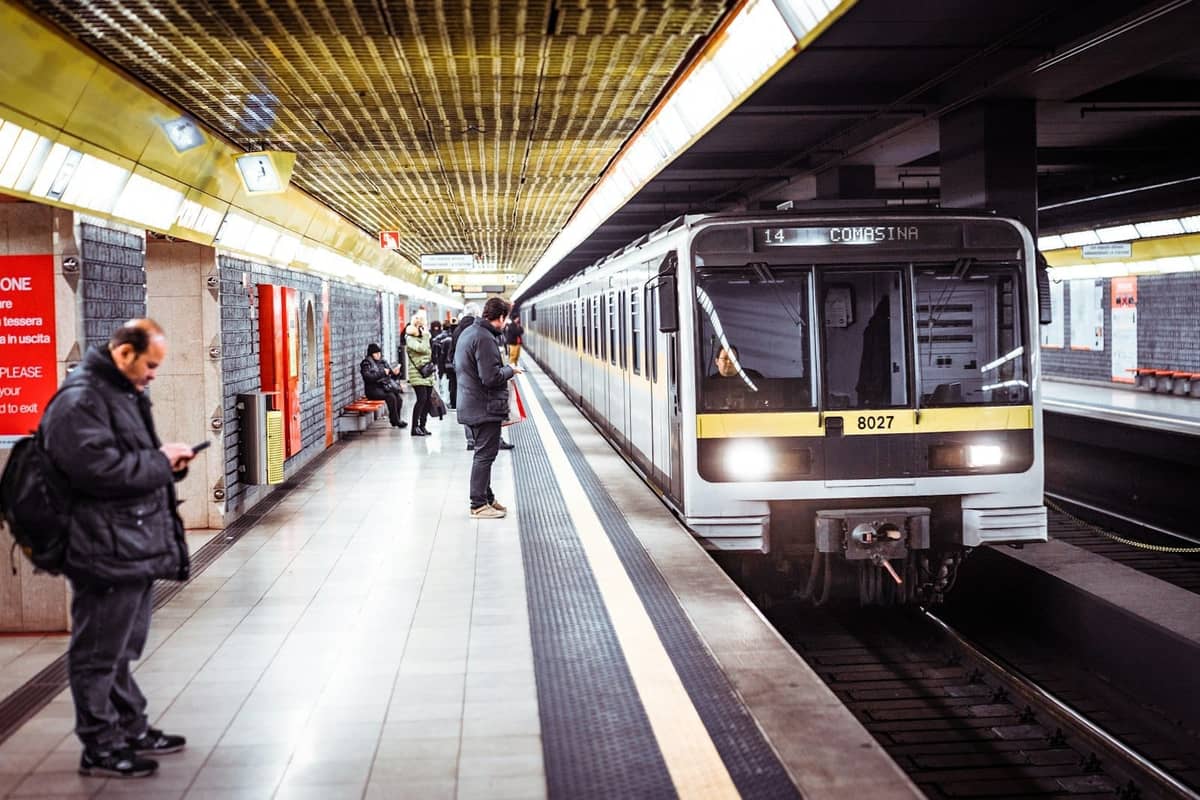 Treno della metropolitana di Milano in arrivo a una banchina sotterranea con persone in attesa e moderne luci gialle sul soffitto.
