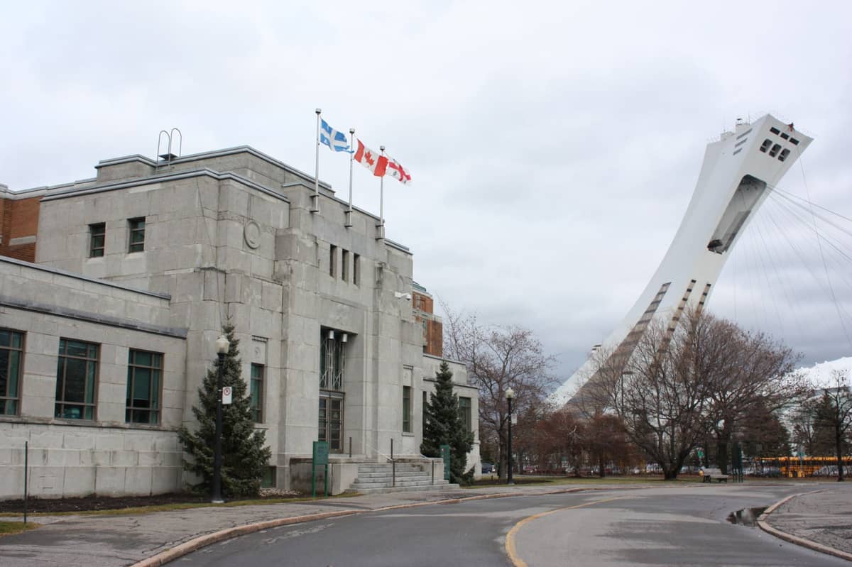 Montreal Olympic Stadium tower rises behind an adjacent building, with Quebec and Canadian flags, on a grey winter day.