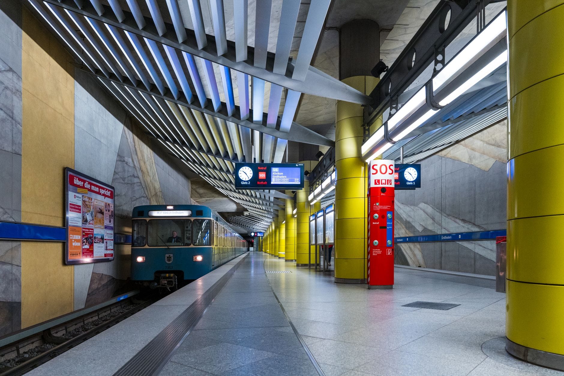 สถานีรถไฟใต้ดิน U-Bahn สมัยใหม่ในมิวนิก มีขบวนรถสีน้ำเงินจอดที่ชานชาลา เสาสีเหลืองสดใส และป้ายบอกทางชัดเจนเหนือราง