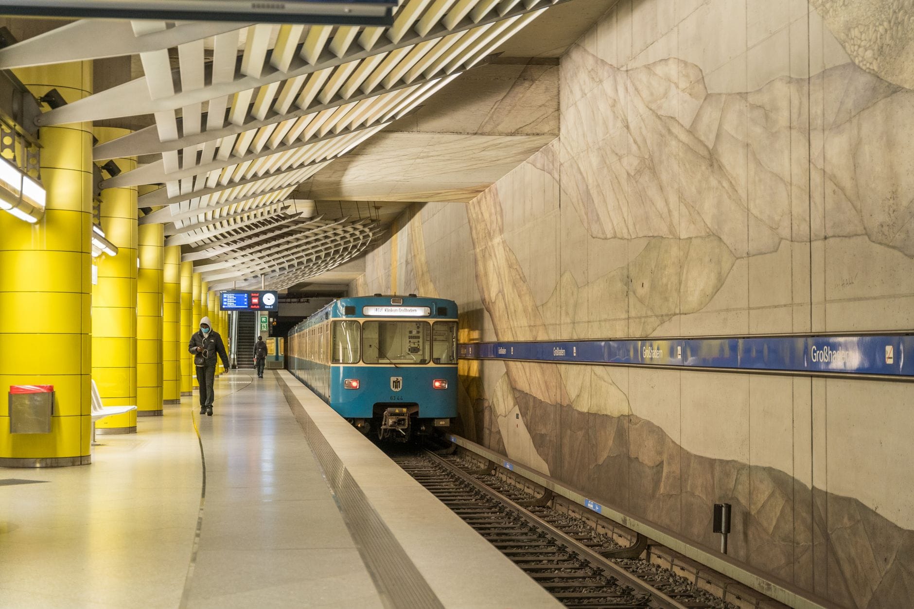สถานี U-Bahn ในมิวนิกพร้อมรถไฟใต้ดินสีน้ำเงิน เสาสีเหลือง และผู้โดยสารบนชานชาลา