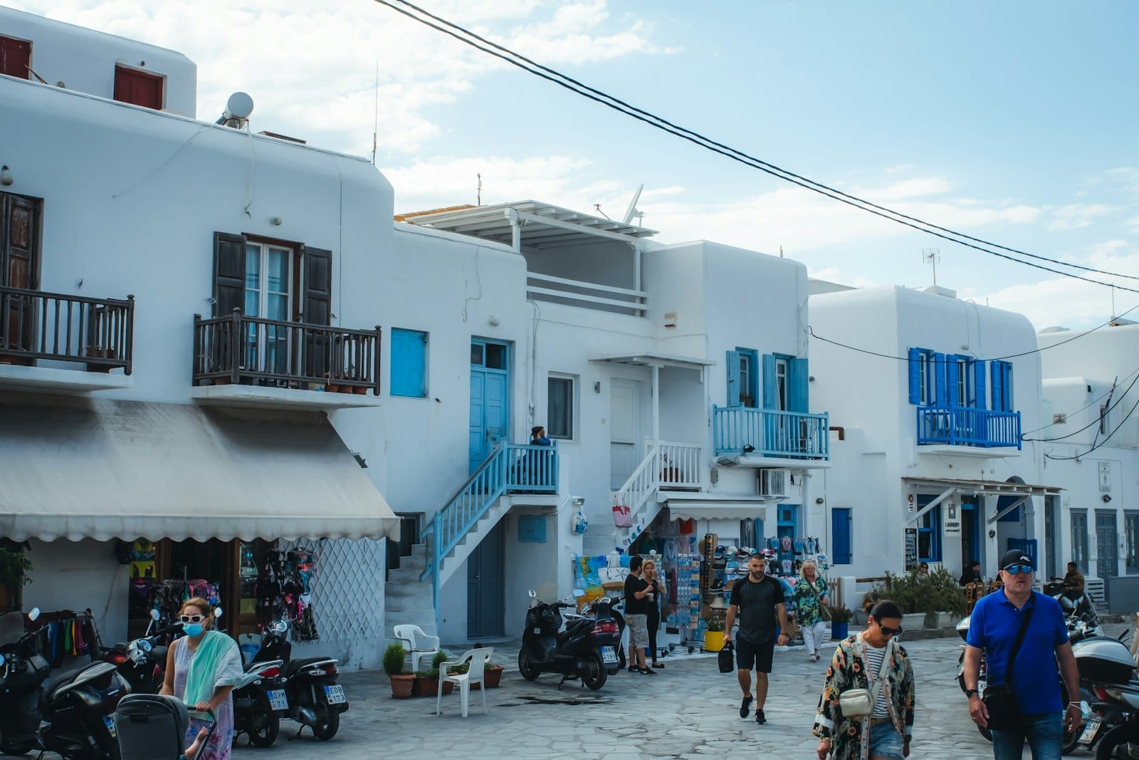 Personas caminando por una calle de Mykonos flanqueada por edificios blancos, con scooters y motocicletas estacionados en primer plano.