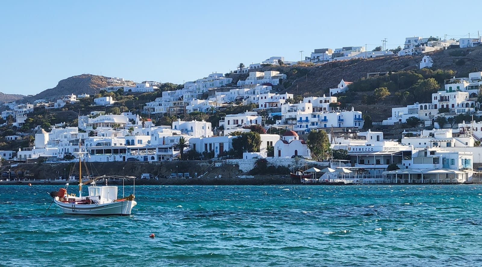 Un pequeño bote navega sobre aguas turquesas frente a una ladera cubierta de edificios cicládicos blancos, capturando el transporte y el paisaje de Mykonos.