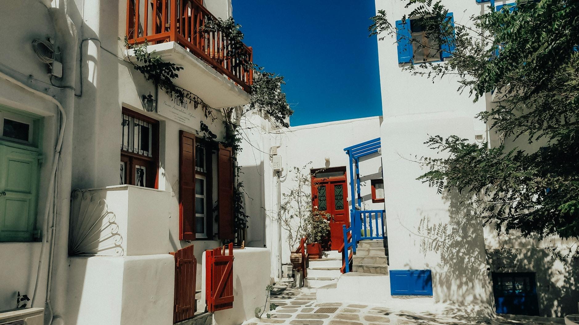 Callejón típico de Mykonos Town con edificios encalados, puertas y contraventanas de colores, estrecho camino de piedra y cielo azul.