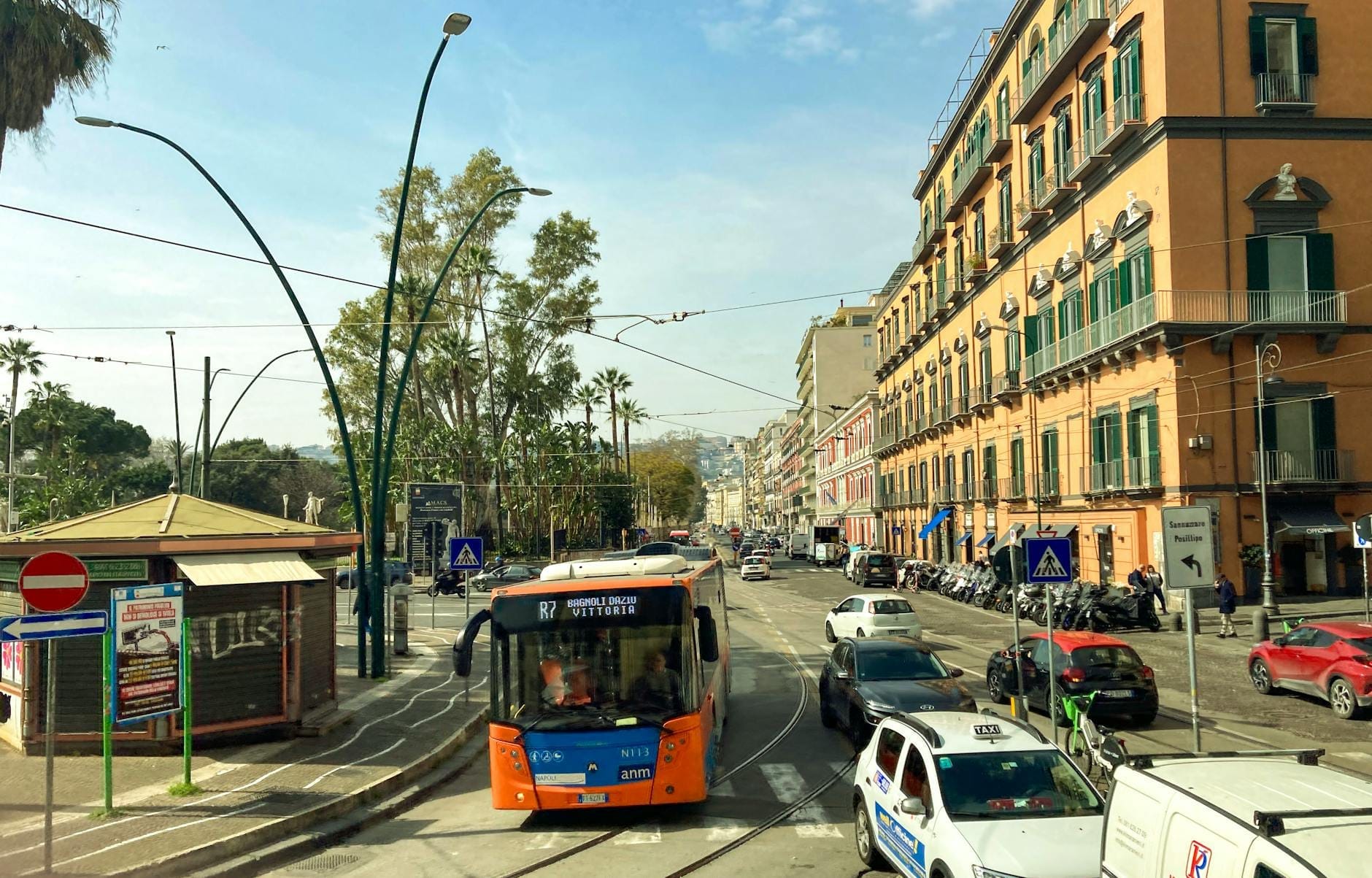 Uma rua de Nápoles com um ônibus urbano laranja da ANM, linhas de bonde, edifícios coloridos e pessoas em um ponto de ônibus sob um céu limpo.