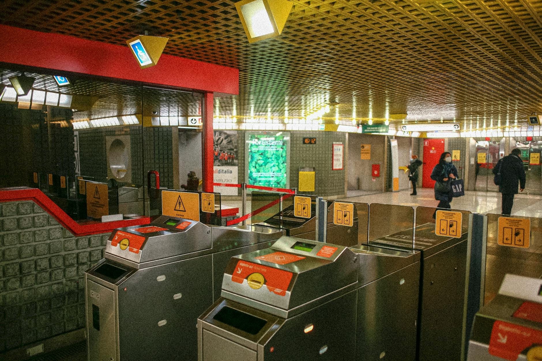 Entrada da estação de metrô de Nápoles com catracas ANM, sinalização colorida e pessoas entrando na área da plataforma.