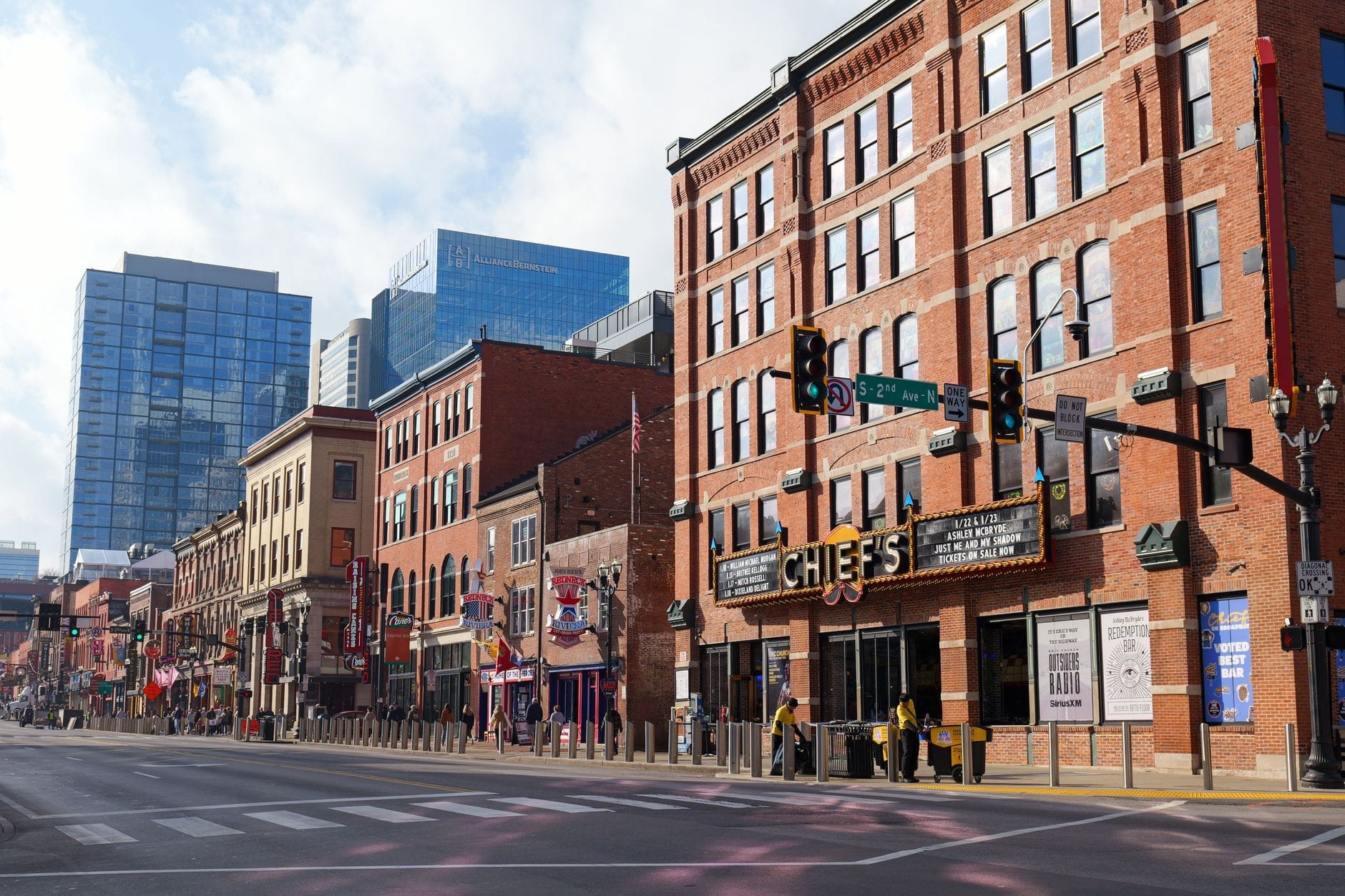 Escena diurna de una calle en el centro de Nashville con históricos edificios de ladrillo, letreros de neón y modernos rascacielos al fondo.