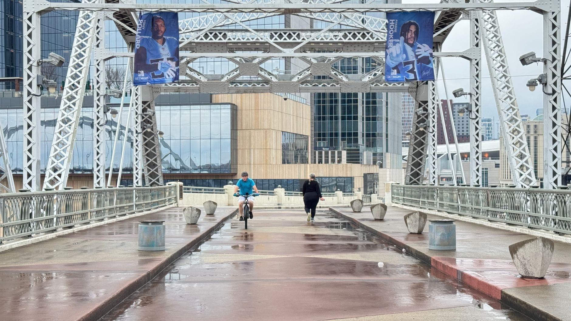 Personas caminando y andando en bicicleta en el Puente Peatonal John Seigenthaler de Nashville con los edificios del centro al fondo.
