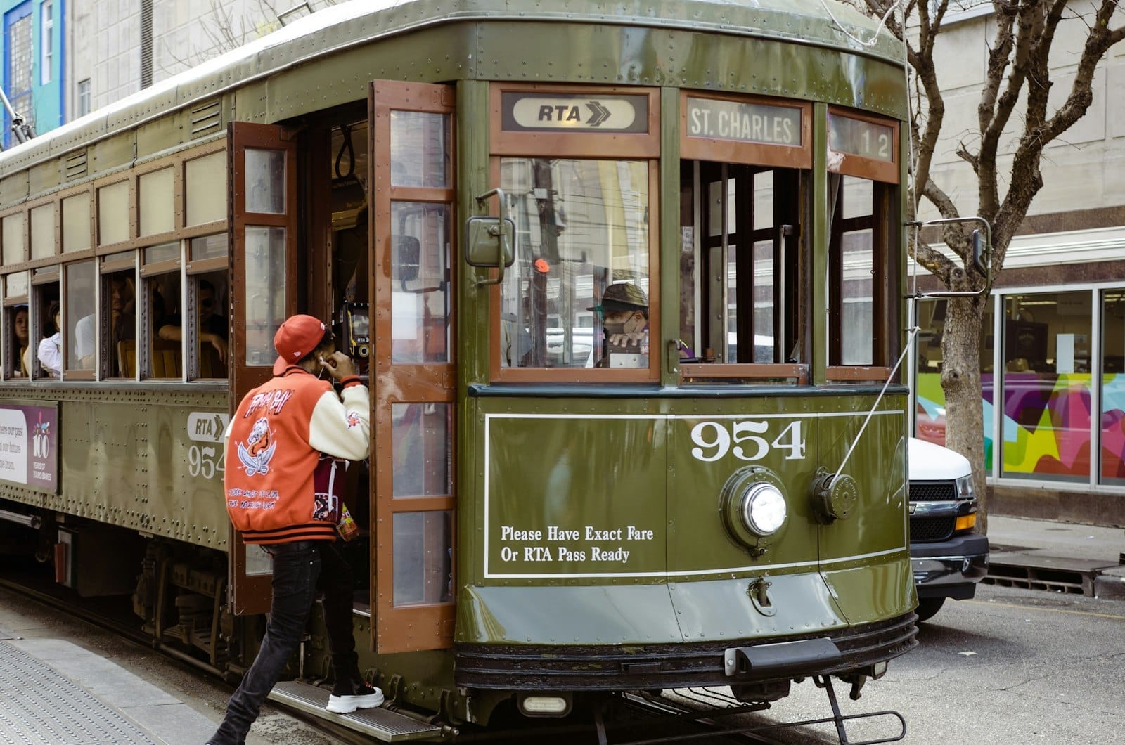 Bonde clássico verde de Nova Orleans com um passageiro embarcando, rua urbana e mural colorido ao fundo, destacando o transporte público histórico da cidade.