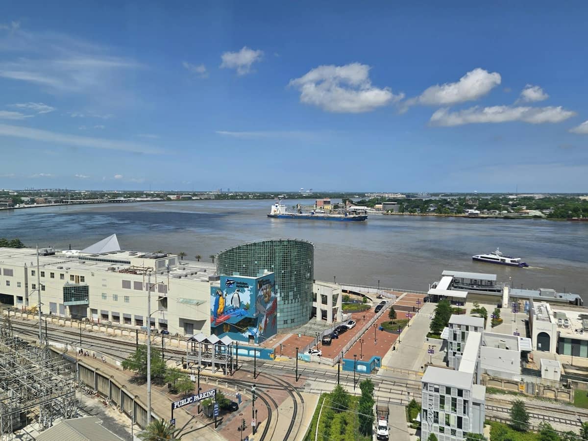 A view of the Mississippi River, New Orleans waterfront, ferries, and streetcar tracks on a clear day with city landmarks visible.
