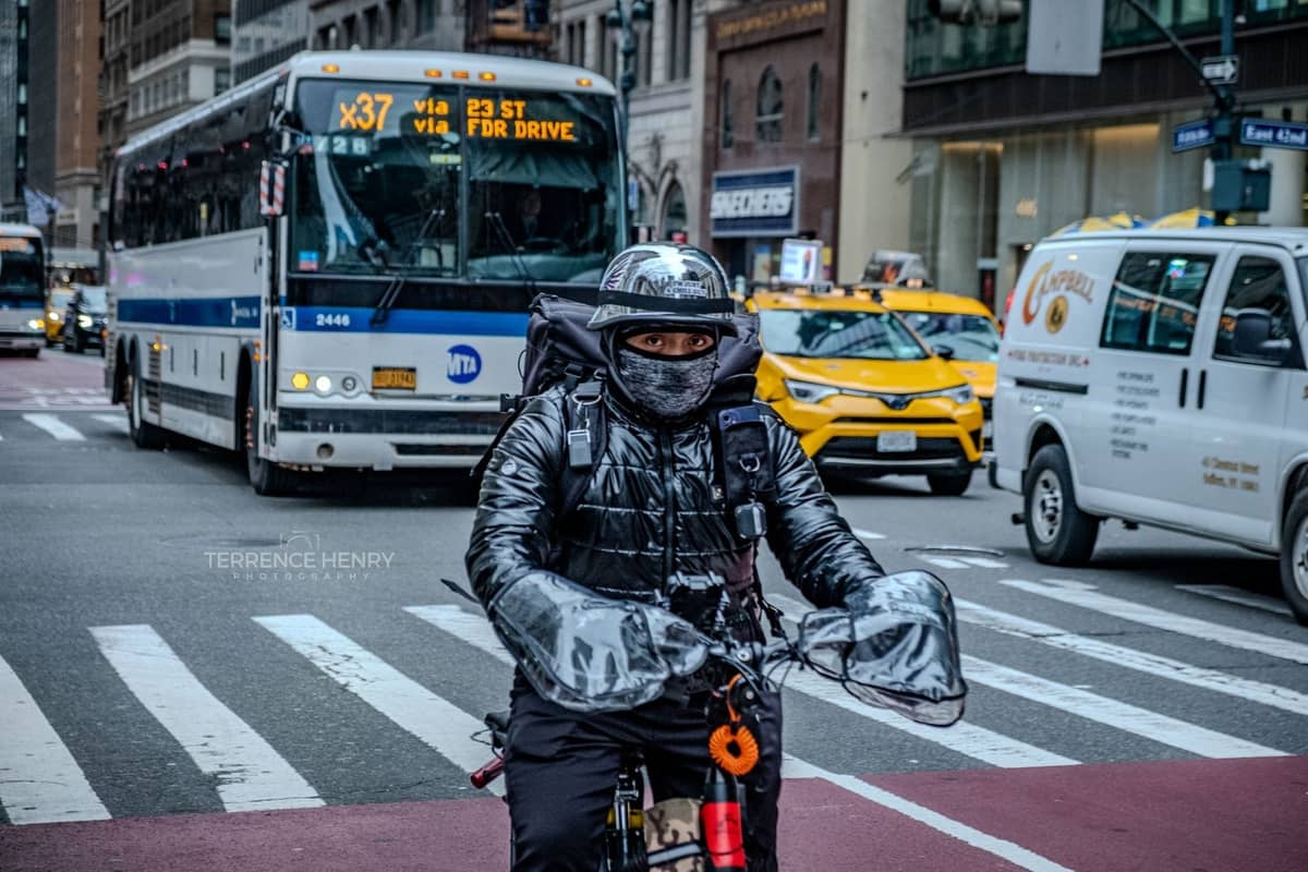 Eine Person auf einem Fahrrad fährt auf einer Straße in New York City, im Hintergrund sind ein MTA-Bus, Taxis und Stadtgebäude zu sehen.