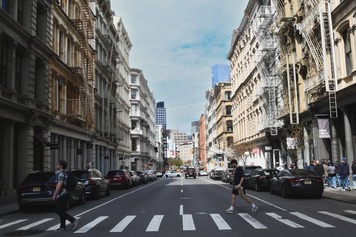 Breite Straßenansicht von New York City mit Menschen, die einen Zebrastreifen überqueren, klassischen Gebäuden und klarem Himmel im Hintergrund.